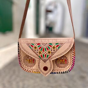 Puede incluir: Bolso bandolera de cuero color beige con correa marrón. El bolso presenta diseños geométricos coloridos e ilustraciones de camellos. Los bordes están cosidos con un arcoíris de colores. La palabra "MAROC" es visible en el bolso.