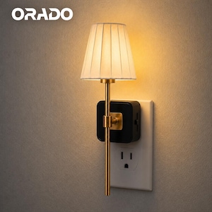 May include: A plug-in wall lamp with a gold-toned metal stem and a pleated white fabric shade. The lamp features a black square base that plugs directly into a standard white wall outlet, emitting a warm, soft glow from the light source.