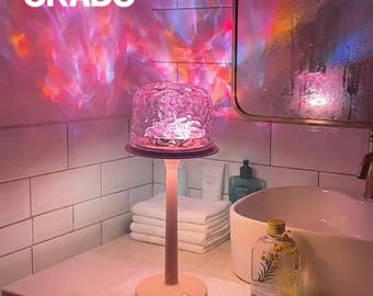 Op de afbeelding: Een roze tafellamp met een heldere, getextureerde kap. De lamp projecteert kleurrijke lichtpatronen op de muur. Het woord "ORADO" staat in witte letters boven de lamp.