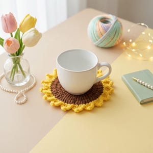 Op de afbeelding: Een witte keramische mok staat op een handgemaakte gehaakte zonnebloemonderzetter. Een kleine glazen vaas bevat tulpen en een bol garen ligt in de buurt. Een notitieboek en een pen liggen ook op tafel.