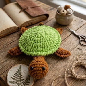 May include: A handmade crochet turtle with a green shell and brown head, flippers, and tail. The turtle is placed on a rustic wooden surface, with a fern-leaf ceramic coaster, open book, and small scissors in the background.