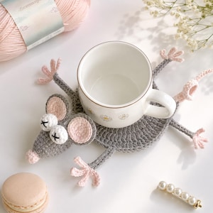 May include: A gray crocheted mouse-shaped coaster with pink accents, holding a white ceramic mug. The coaster has large pink ears and white pom-pom eyes. A ball of pink yarn and a pearl hair clip are also visible.