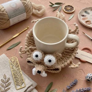 Könnte beinhalten: Eine beige Keramiktasse steht auf einem handgefertigten, gehäkelten Untersetzer in Form einer Cartoon-Kreatur mit großen weißen Augen und einer weißen Schnauze. Der Untersetzer ist beige und hat ein Etikett mit der Aufschrift "HANDMADE".