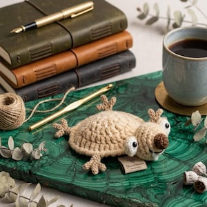 May include: A handmade crochet sloth toy in tan with white and brown accents, lying on a green marble surface. A gold crochet hook, a ball of twine, and a stack of leather-bound journals are also visible.