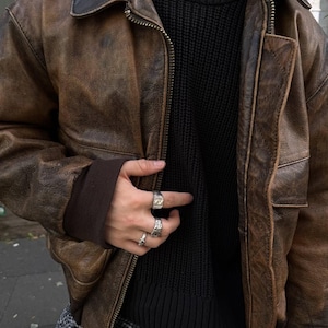 Op de afbeelding: Een bruine leren bomberjack met een zwarte gebreide trui eronder. De jas heeft een ritssluiting en twee zakken aan de voorkant. De persoon draagt ​​meerdere zilveren ringen aan zijn vingers.
