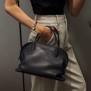 May include: A black leather handbag with a rounded shape and two top handles. The bag has a textured surface and a zipper closure. The person is wearing a black top, light-colored pants, and a gold watch.