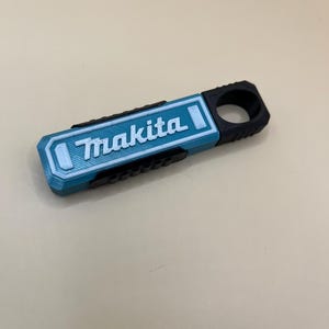 May include: A 3D-printed tool with the "Makita" logo in white on a teal rectangular base. It has a black handle and textured sides. The design is modern and looks like a custom accessory. The tool is likely for DIY or construction.