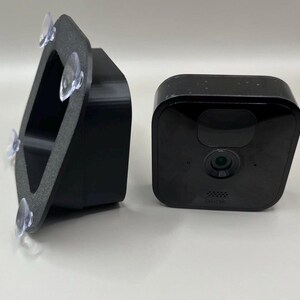 May include: A black square security camera with a central lens and the word blink printed below it, positioned next to a black mounting bracket featuring four clear suction cups attached to its textured, angled frame for window mounting.