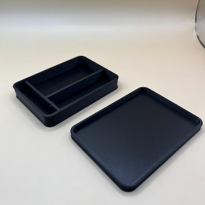 May include: A dark navy blue rectangular tray with a lid. The tray is divided into three compartments. The lid is a flat, solid rectangle. The items are made of a matte material and sit on a white surface.