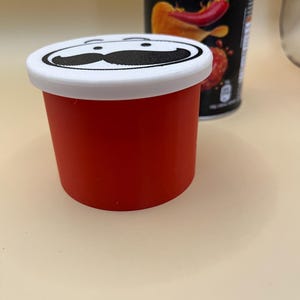 May include: A red container with a white lid and a black mustache design. Behind it is a black can with the text "Blazin' Fried Chicken Flavour" and a chili pepper graphic.