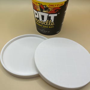 May include: A white Pot Noodle cup with a black and yellow label that reads "Pot Noodle Bombay Bad Boy". Two white lids are in front of the cup. The cup is on a light tan surface.