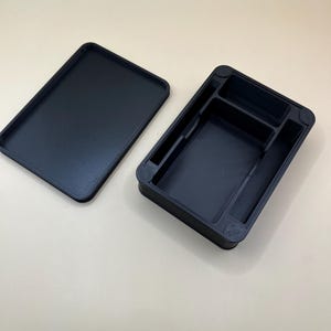 May include: A black rectangular storage box with a separate lid. The open box has several compartments of different sizes. The lid is placed next to the box. Both the box and lid are made of a matte black material.