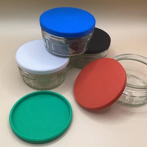 May include: Four glass jars with lids. The lids are in different colours: blue, white, black, and red. The jars are clear glass and have a textured surface.