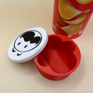 May include: A red and white container with a cartoon face lid. The container is open, showing a red interior. A red Pringles tube is in the background with chips visible.