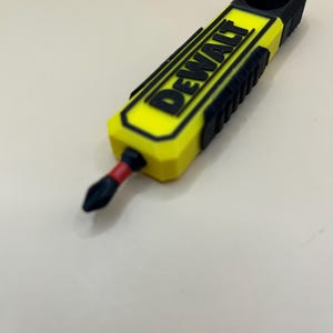 May include: A yellow and black screwdriver with the word "DEWALT" in black lettering. The screwdriver bit is black and red. The tool is on a white surface.