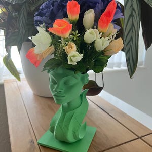 May include: A green head-shaped vase filled with artificial flowers in shades of orange, white, and peach, with blue and green accents. The vase sits on a square base. The arrangement is displayed on a wooden surface.
