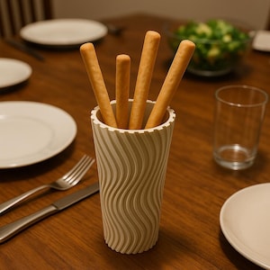 May include: A beige, textured breadstick holder filled with breadsticks. The holder sits on a wooden table, alongside plates, cutlery, and a glass. A bowl of salad is visible in the background.