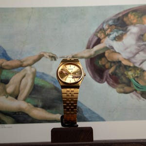 May include: A gold-toned Casio watch with a matching band is displayed in front of a print of Michelangelo's "The Creation of Adam." The watch has a gold face and a rectangular case. The background features the iconic fresco.