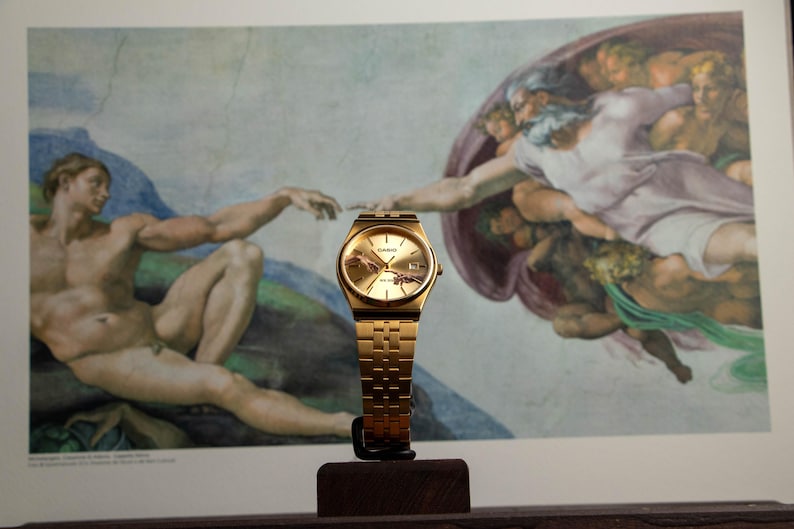 May include: A gold-toned Casio watch with a matching band is displayed in front of a print of Michelangelo's "The Creation of Adam." The watch has a gold face and a rectangular case. The background features the iconic fresco.