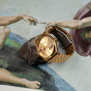 May include: Gold-toned Casio watch with a rectangular face and a gold link band, resting on a dark rock. The watch face features a depiction of the 'Creation of Adam' artwork. The background is a mural with figures.