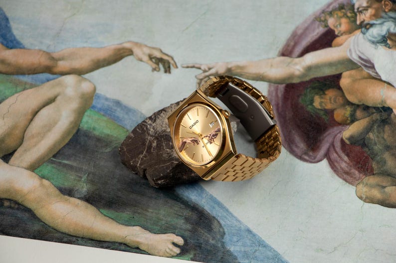 May include: Gold-toned Casio watch with a rectangular face and a gold link band, resting on a dark rock. The watch face features a depiction of the 'Creation of Adam' artwork. The background is a mural with figures.