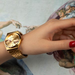 May include: Gold-toned wristwatch with a rectangular face and matching band. The watch face features a detailed design. The watch is worn on a wrist with red nail polish. The background is a painting.