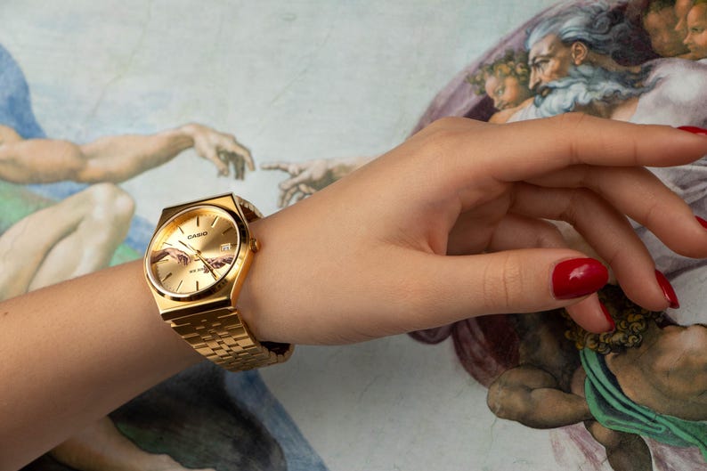 May include: Gold-toned wristwatch with a rectangular face and matching band. The watch face features a detailed design. The watch is worn on a wrist with red nail polish. The background is a painting.