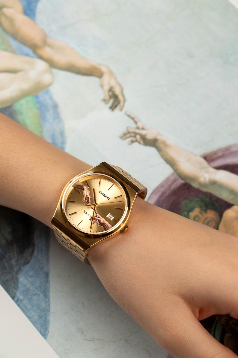 May include: Gold-toned Casio watch with a rectangular face and a gold band. The watch face features the iconic 'Creation of Adam' artwork. The watch is displayed on a wrist against a backdrop of the same artwork.