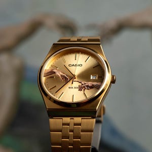 May include: Gold-toned Casio watch with a rectangular face and matching band. The watch face features the iconic 'Creation of Adam' artwork. The watch is a classic design with a modern twist, suitable for everyday wear.