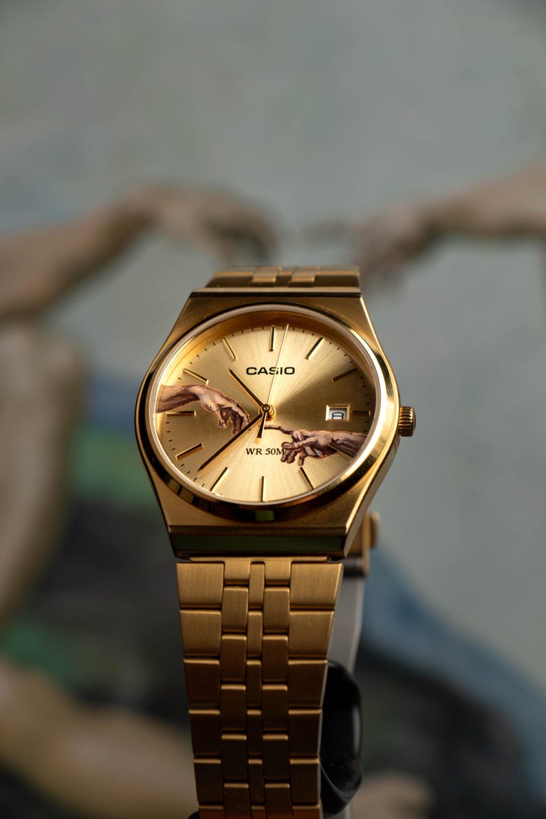 May include: Gold-toned Casio watch with a rectangular face and matching band. The watch face features the iconic 'Creation of Adam' artwork. The watch is a classic design with a modern twist, suitable for everyday wear.
