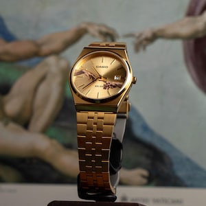 May include: Gold-toned Casio watch with a rectangular face and a matching link band. The watch face features the iconic "Creation of Adam" artwork. The watch is displayed on a dark wooden stand, showcasing its elegant design.