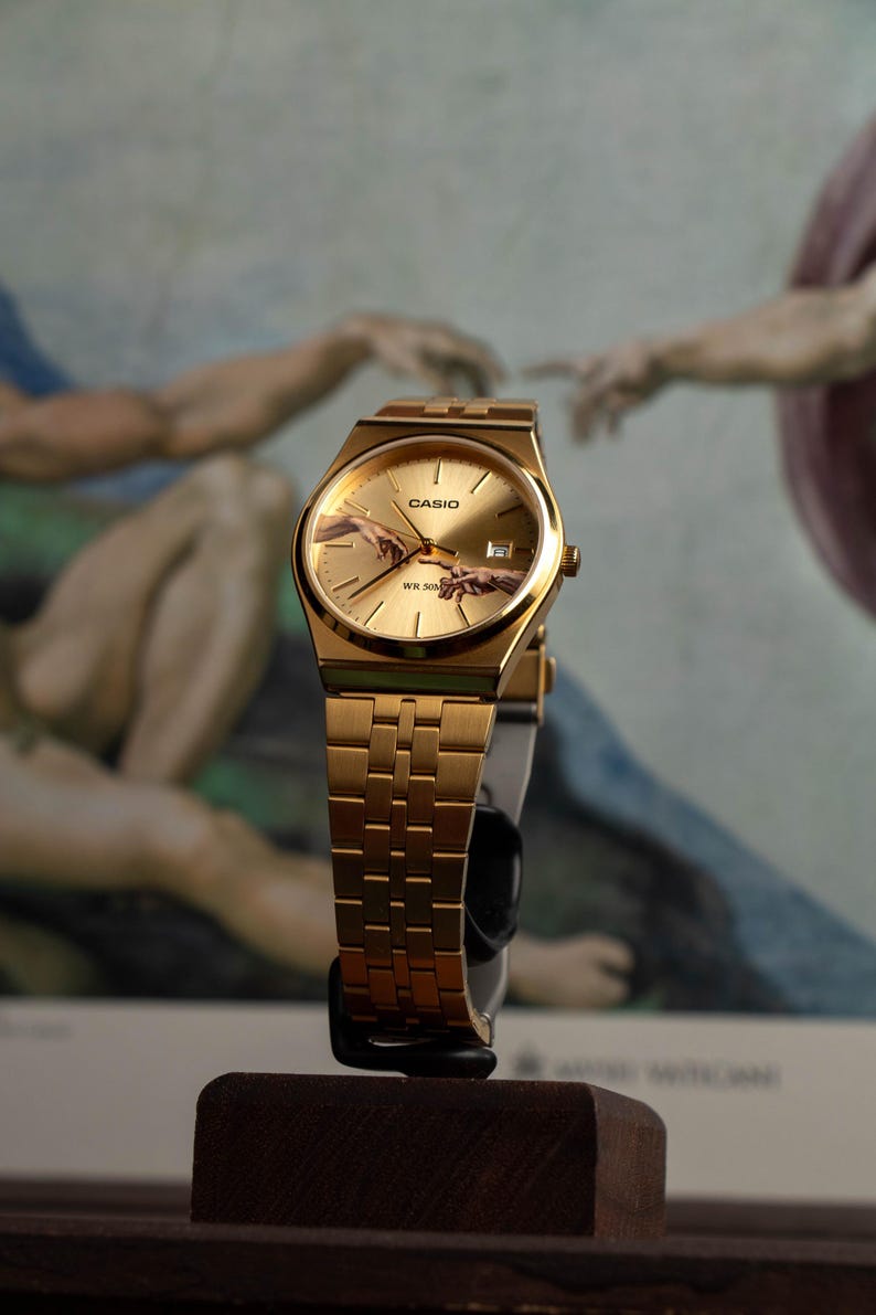 May include: Gold-toned Casio watch with a rectangular face and a matching link band. The watch face features the iconic "Creation of Adam" artwork. The watch is displayed on a dark wooden stand, showcasing its elegant design.