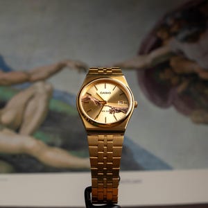 May include: Gold-toned Casio watch with a rectangular face and matching band. The watch face features an image of the Sistine Chapel's "Creation of Adam" painting. The watch is displayed against a backdrop of the same artwork.