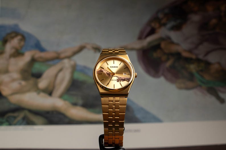 May include: Gold-toned Casio watch with a rectangular face and matching band. The watch face features an image of the Sistine Chapel's "Creation of Adam" painting. The watch is displayed against a backdrop of the same artwork.