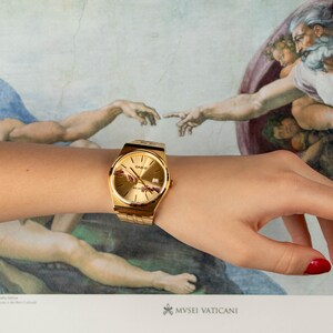 May include: Gold-toned wristwatch with a rectangular face and a matching band. The watch is worn on a wrist, with a background featuring a detail from the Sistine Chapel ceiling. The watch face has a depiction of the Creation of Adam.