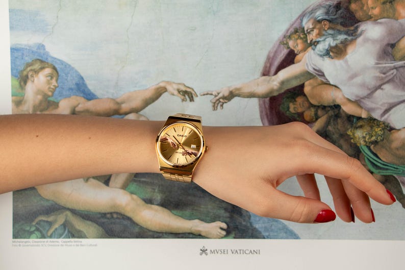 May include: Gold-toned wristwatch with a rectangular face and a matching band. The watch is worn on a wrist, with a background featuring a detail from the Sistine Chapel ceiling. The watch face has a depiction of the Creation of Adam.