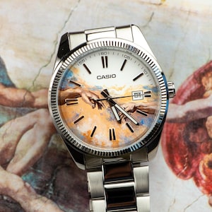 May include: A silver Casio watch with a white face featuring a painting of two hands reaching out. The watch has a silver band and a date display. The background shows a portion of a Renaissance painting.