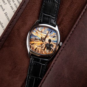 May include: A wristwatch with a silver-toned case and a black leather strap featuring a textured crocodile pattern. The watch face displays a detailed, colourful painted scene of a person standing in a golden wheat field under a cloudy sky.