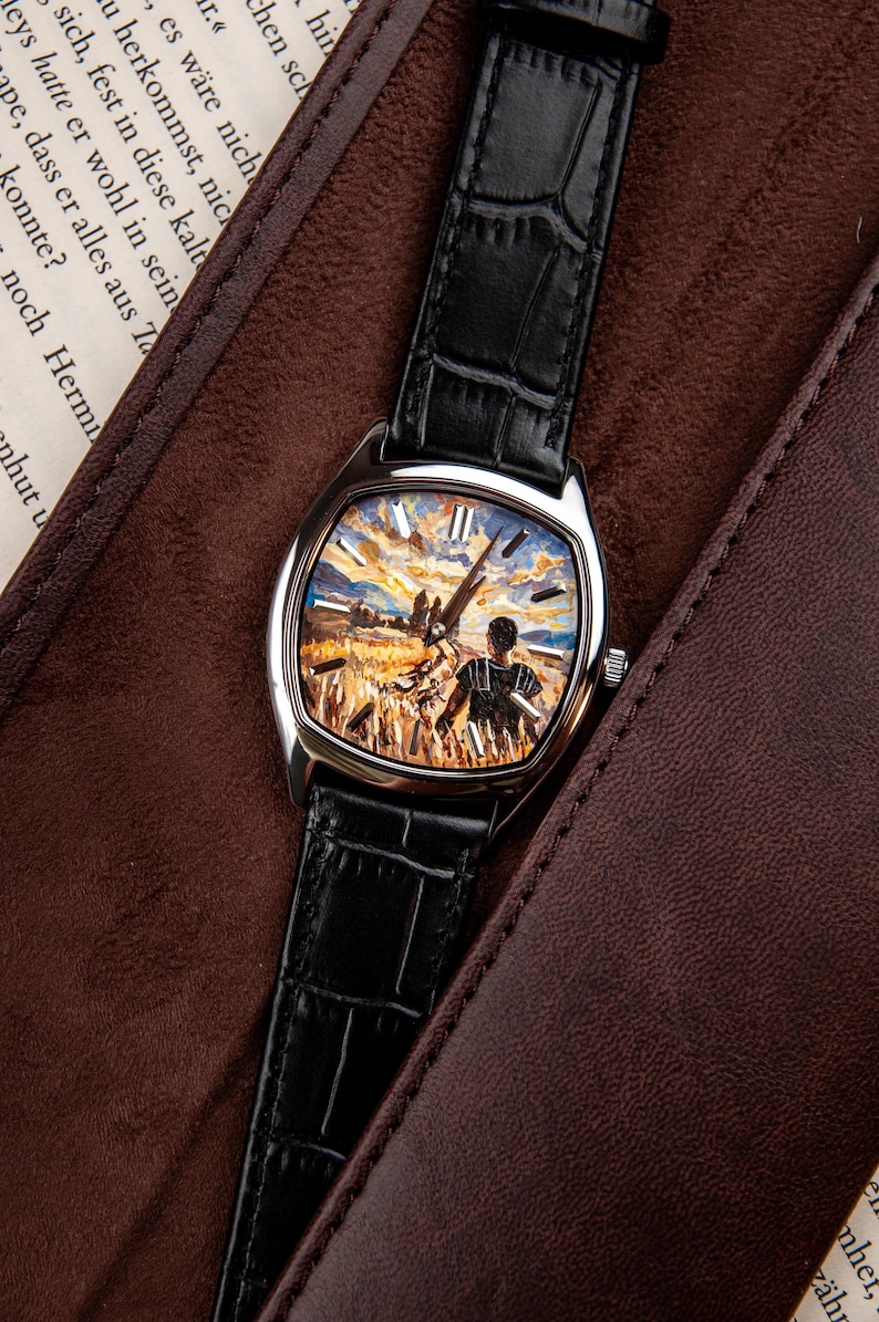 May include: A wristwatch with a silver-toned case and a black leather strap featuring a textured crocodile pattern. The watch face displays a detailed, colourful painted scene of a person standing in a golden wheat field under a cloudy sky.