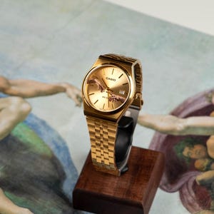 May include: Gold-toned wristwatch with a rectangular face and a matching link band. The watch face features a depiction of the 'Creation of Adam' from the Sistine Chapel. The watch is displayed on a small wooden stand.