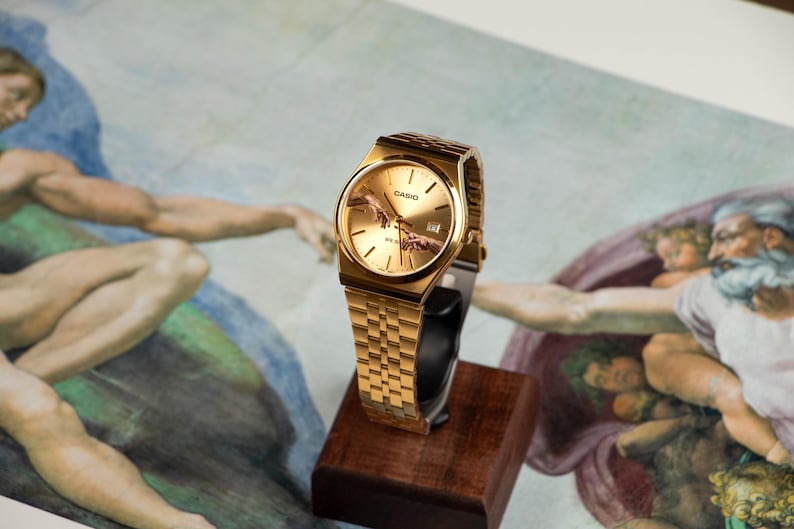 May include: Gold-toned wristwatch with a rectangular face and a matching link band. The watch face features a depiction of the 'Creation of Adam' from the Sistine Chapel. The watch is displayed on a small wooden stand.