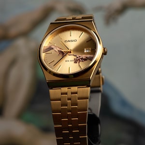 May include: Gold-toned Casio watch with a rectangular face and matching band. The watch face features the iconic 'Creation of Adam' artwork. The watch has the Casio logo and 'WR 50M' text. The watch is displayed against a blurred background.