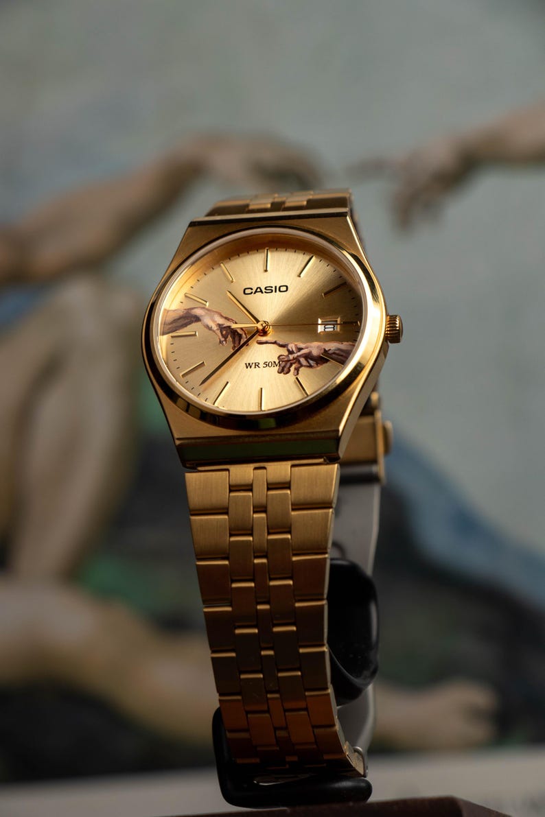 May include: Gold-toned Casio watch with a rectangular face and matching band. The watch face features the iconic 'Creation of Adam' artwork. The watch has the Casio logo and 'WR 50M' text. The watch is displayed against a blurred background.