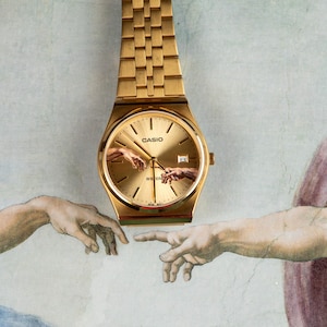 May include: Gold-toned Casio watch with a gold dial and band. The watch face features an artistic depiction of two hands reaching out, inspired by the Sistine Chapel. The watch is set against a light-colored background.