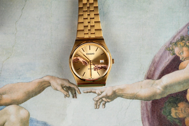 May include: Gold-toned Casio watch with a gold dial and band. The watch face features an artistic depiction of two hands reaching out, inspired by the Sistine Chapel. The watch is set against a light-colored background.