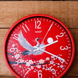 Puede incluir: Reloj de pared redondo rojo con borde negro. La esfera del reloj presenta un diseño de grulla y flor de cerezo. Agujas y números blancos visibles. El reloj está montado sobre una superficie de madera.
