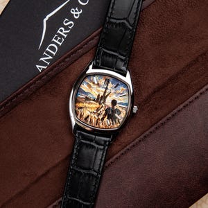 May include: A wristwatch with a black crocodile-embossed leather strap and a silver-tone case. The watch face features a detailed painted scene of a person standing in a golden wheat field under a sunset sky. The brand name ANDERS & CO is visible.