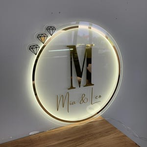 May include: A round, illuminated sign with a gold border and the text "Mia & Leo" in gold script. The letter "M" is prominently displayed in gold. Three diamond graphics are on the left side of the sign.