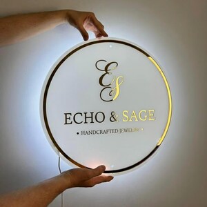 May include: A round, illuminated sign with a white face and gold trim. The sign features the text "ECHO & SAGE" in gold lettering, along with the words "HANDCRAFTED JEWELRY". The sign is held by two hands.