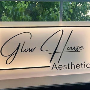 May include: A rectangular illuminated sign with the words "Glow House Aesthetics" in a stylish script font. The sign has a white background with a black border and is likely used for a business.
